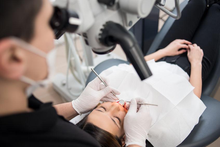 Kobieta na fotelu dentystyczną z otwartą buzią, podczas zabiegu. Nad nia stoi dentysta i patrzy w mikroskop stomatologiczny.