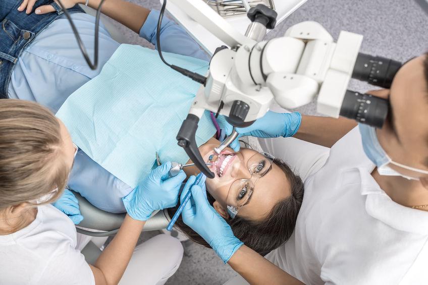 Uśmiechnięta pacjentka na fotelu dentystycznym. Nad nia stoi lekarz i patrzy w mikroskop a obok stoi kobieta, pomoc dentystyczna.